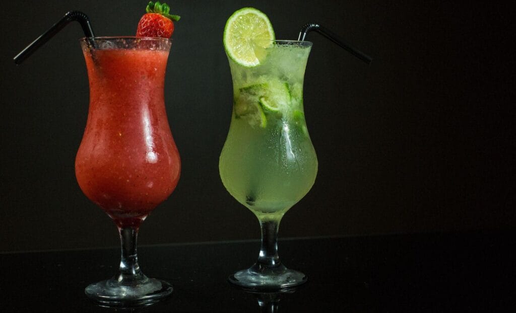 Two brightly coloured cocktails in hurricane glasses with garnishes
